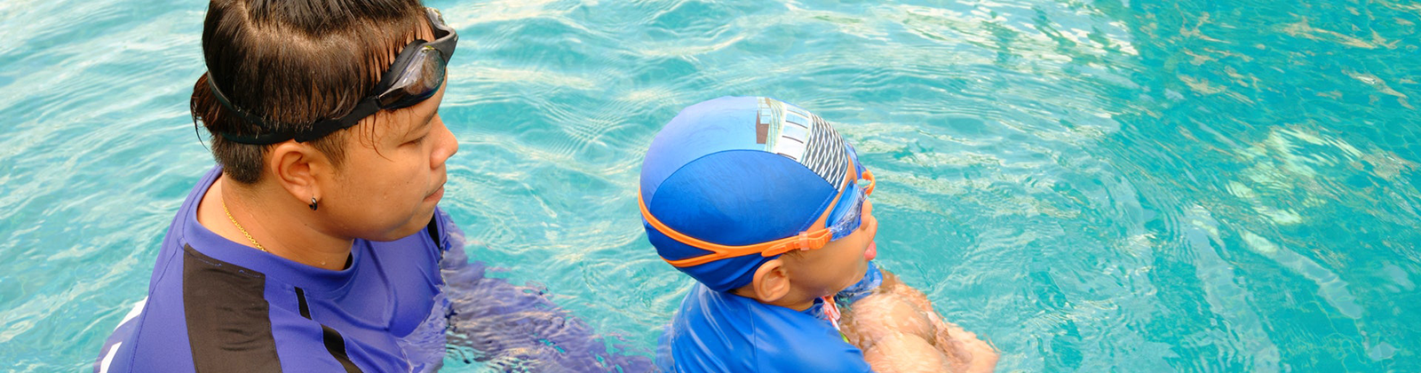 An adult is holding a child in the water. The child is deeply inhaling before going under the water and they are wearing goggles and a cap.